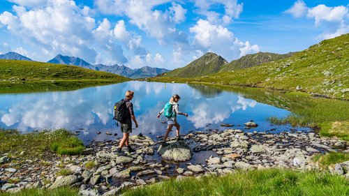 Camminare, pedalare, e sciare in Tirolo Due escursionisti attraversano un lago roccioso in montagna con cielo limpido