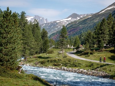 Unsere Tipps für einen Wanderurlaub in Osttirol Zwei Radfahrer auf einem Weg neben einem Fluss und Tannen in den Bergen mit Schnee