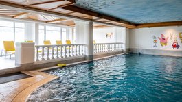 I servizi del nostro hotel per famiglie in Tirolo Piscina coperta con getti d'acqua e sedie gialle vicino a murales di cartoni animati