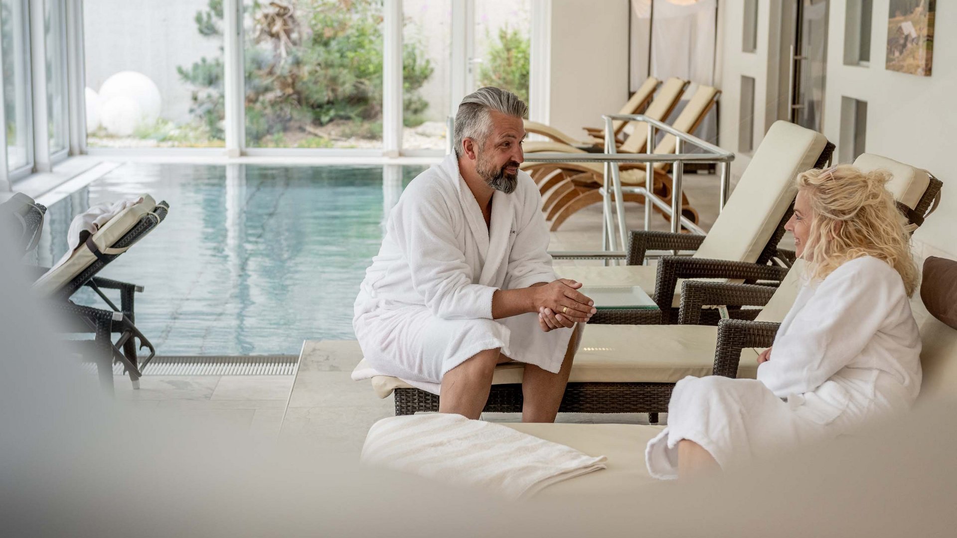 Wellnesshotel im Defereggental: Wohlfühlurlaub vom Feinsten Mann und Frau in Bademänteln neben einem Innenpool auf Liegestühlen