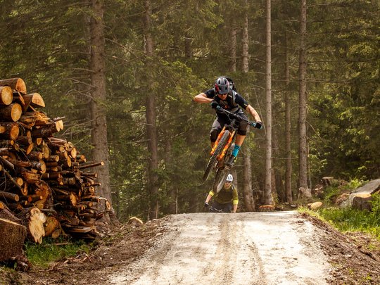 Mountain biking routes for all levels Mountain biker jumps on forest trail over dirt mound with two cyclists behind