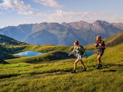 Wandern in Österreich – der Berg ruft! Zwei Wanderer mit Rucksäcken auf einer Bergwiese vor Seen und Berggipfeln