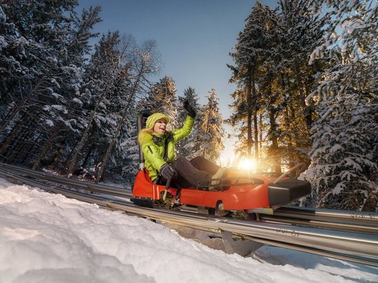 Family hotel in East Tyrol: the Jesacherhof Woman riding bobsled on alpine coaster in snowy forest at sunset