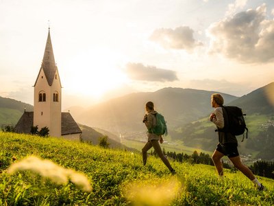 Trekking in Tirolo, Austria: allo Jesacherhof Due escursionisti con zaini camminano su prato verde con chiesa e montagne sullo sfondo