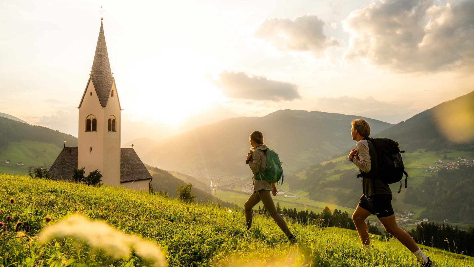 Wandern in Österreich – der Berg ruft! Zwei Wanderer mit Rucksäcken wandern auf grüner Wiese bei malerischer Bergkulisse und Kirche