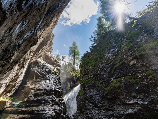 Defereggental: Sehenswürdigkeiten und Ausflugsziele Person überquert eine Hängebrücke über eine Schlucht mit Wasserfall bei Sonnenschein