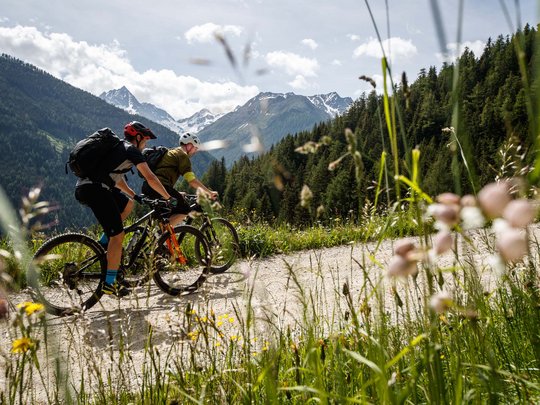 Mountain biking routes for all levels Two mountain bikers riding on a trail with snow-capped mountains in the background