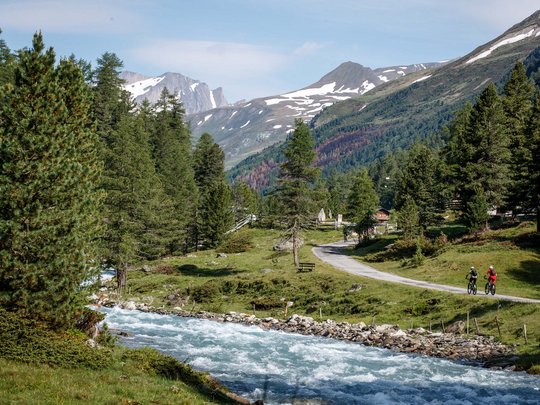 Mountain biking routes for all levels Two cyclists on a path by a river in the mountains with pine trees and snow