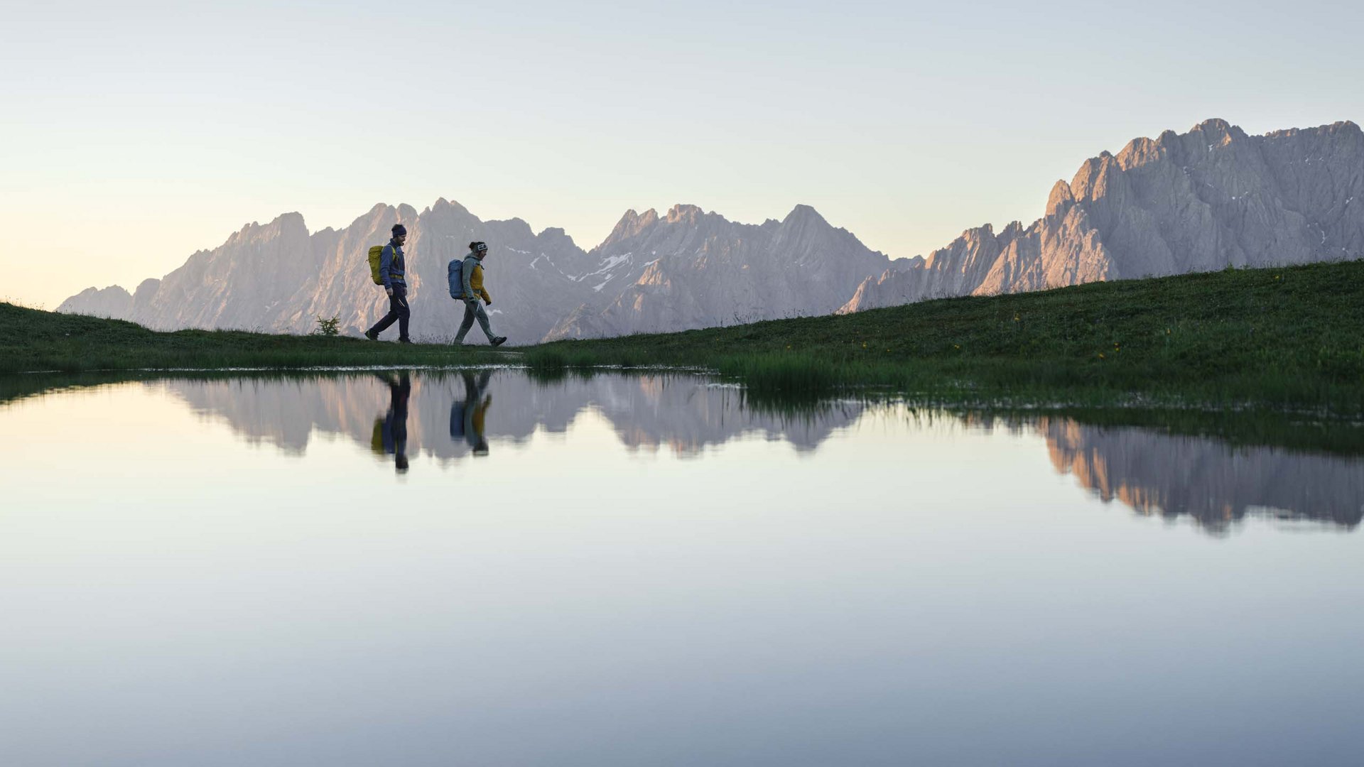 Unvergesslicher Skiurlaub im Defereggental Zwei Wanderer gehen am Bergsee mit Alpen im Hintergrund bei Sonnenuntergang