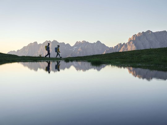 Ausflugsziele im Defereggental Zwei Wanderer gehen am Bergsee mit Alpen im Hintergrund bei Sonnenuntergang