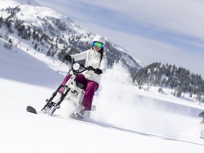 Unvergesslicher Skiurlaub im Defereggental Frau fährt Snowbike mit weißem Helm und pinker Hose im Schnee vor Bergkulisse