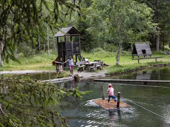 Defereggental: Sehenswürdigkeiten und Ausflugsziele Kind auf einer Holzbarke auf einem kleinen See, Erwachsene am Ufer im Waldpark
