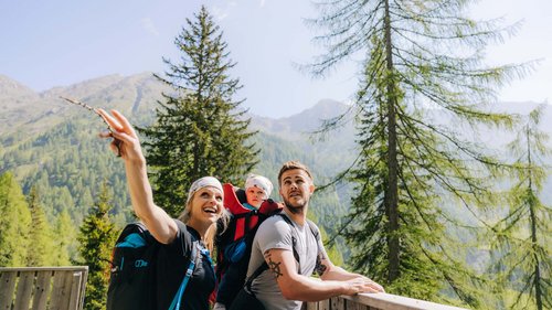 Camminare, pedalare, e sciare in Tirolo Famiglia con bambino in zaino da trekking in montagna con pini e paesaggio montano